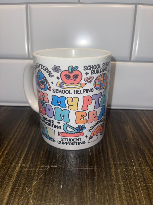 White ceramic mug featuring a colorful design with the words “In My PTA Mom Era” surrounded by playful icons representing volunteer work, teacher support, and school activities.